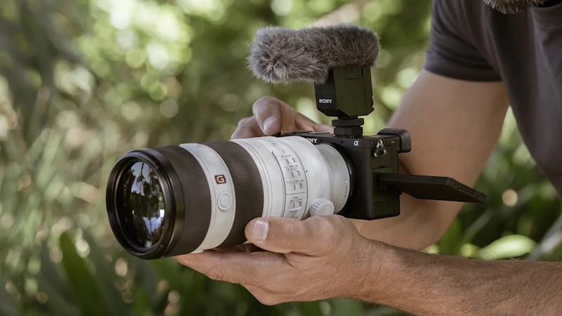 Камера Sony Alpha 6700 с открытым поворотным экраном, установленным микрофоном Sony с ветрозащитой и объективом в руках оператора на фоне парка
